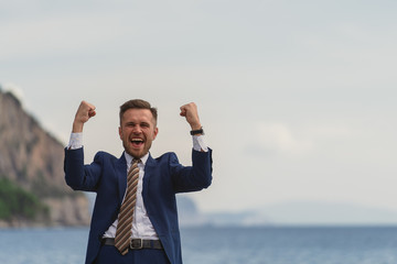 Excited businessman in a suit screaming and raising hands to the sky by the sea