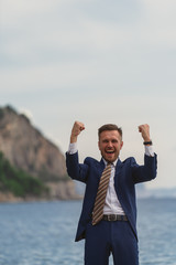 Excited businessman in a suit screaming and raising hands to the sky by the sea