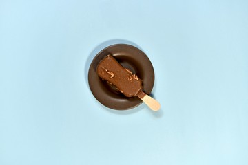 Chocolate ice-cream with nut, on a stick. In a saucer, on a blue background, in the center. View from above.