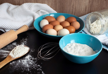 Preparation of the dough. Flour, eggs, whisk, rolling pin and a kitchen towel on the black table. A bunch of flour on a wooden spoon, a bowl of flour and lots of eggs on the blue plate. Baking reciepe