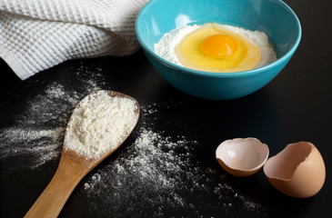 Flour, egg and a kitchen towel. Preparation of the dough on dark background.  A bunch of flour on a wooden spoon, a broken egg in a bowl of flour and egg shell. Flour scattered on the black table
