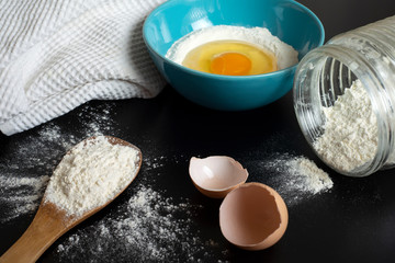 Flour, egg and a kitchen towel. Preparation of the dough on dark background.  A bunch of flour on a wooden spoon, a broken egg in a bowl of flour and egg shell. Flour scattered on the black table