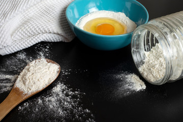 Flour, eggs and a kitchen towel. Preparation of the dough on dark background.  A bunch of flour on a wooden spoon, a broken egg in a bowl of flour. Baking recipe. Flour scattered on the black table