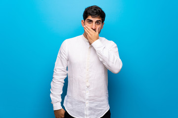 Young man over isolated blue wall covering mouth with hands for saying something inappropriate