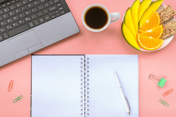 Healthy breakfast at work: cup of black coffee, mango and orange slices and sesame bar on pink background. Notebook, pen, paper clips. Flat lay, copy space.