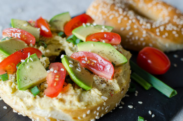 Bagel with hummus, avocado slices, spices, green onion, tomatoes cherry and sesame seeds on the black stone desk. Close up.