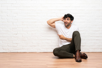 Young man sitting on the floor surprised and pointing finger to the side