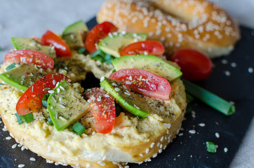 Bagel with hummus, avocado slices, spices, green onion, tomatoes cherry and sesame seeds on the black stone desk. Close up.