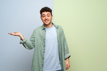 Young man over blue and green background holding copyspace imaginary on the palm to insert an ad
