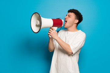 Young man over blue background shouting through a megaphone
