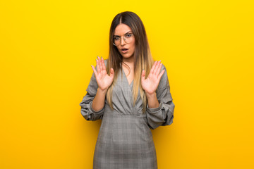 Young woman with glasses over yellow wall making stop gesture with both hands