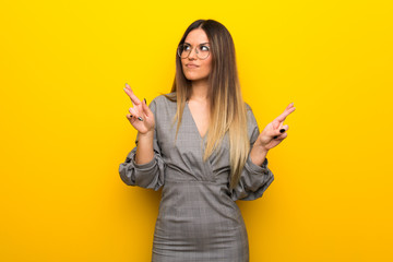 Young woman with glasses over yellow wall with fingers crossing and wishing the best