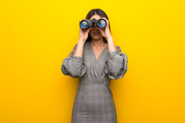 Young woman with glasses over yellow wall and looking in the distance with binoculars