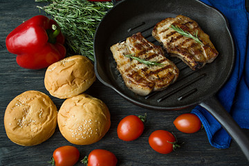 Grilled steak on a round grill pan, garnished with spices for meat, rosemary, greens and vegetables on a dark wooden background.