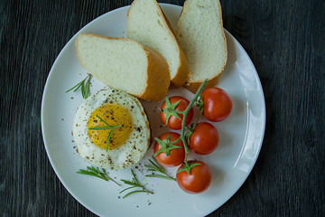 Beef steak with fried egg in spices. Decorated with rosemary, fresh cherry and slices of bread. Filed on a white plate. Dark wooden background. View from above.