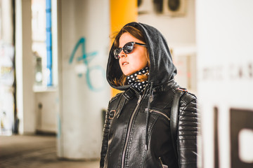 Naklejka premium Young woman with brown hair wearing a leather jacket with a grey hood, a scarf with white dots and black sunglasses – Girl smiling on a bright day outdoor