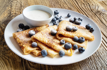 a plate of crepes with sour cream, blueberries, and sugar powder on wooden background