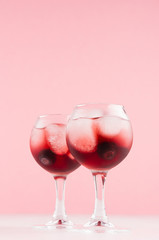 Fresh red berry cocktail with ice cubes, blueberry in elegant misted goblets on soft light pink color background and white wooden table, vertical.