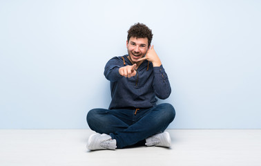 Young man sitting on the floor making phone gesture and pointing front