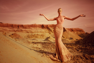 Fototapeta na wymiar Fashionable young blonde woman in the desert in long gold dress stand with open hands, during at sunset background.