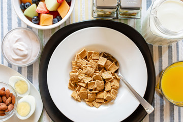 A bowl of cereal as part of a balanced breakfast.  Includes:  Yogurt, almonds, hard boiled egg, mixed fruit, orange juice, and milk.  Flat lay, over head shot.  Background of a striped place mat.