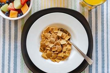 Flat lay of a bowl of cereal with a bowl of mixed fruit in the upper left corner and a glass of orange juice in upper right.  Background of striped multicolored place mat.