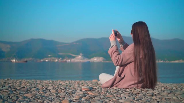 Overall plan. A young girl with long hair in glasses and a coat on the seashore in the spring. A girl makes a photo on the phone. Background sky, mountains and sea.