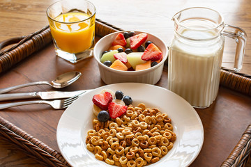 A bowl of oat cereal with fruit and milk on a wooden tray on a wood table.  A bowl of mixed fruit in background with a glass of orange juice and a pitcher of milk.