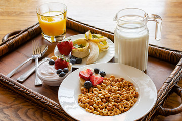 A bowl of oat cereal on wooden tray with assorted fruit, yogurt, orange juice and milk.  Tray on wooden table.  Angled side view.