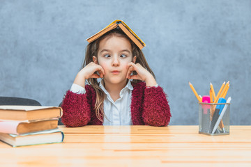Pretty curly girl sitting and holding a book over her head over a grey background. During this schoolgirl takes her eyes away. Stationery products on the table.