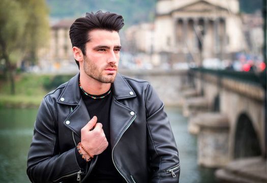 One handsome young man in urban setting in modern city, standing, wearing black leather jacket and jeans, looking away