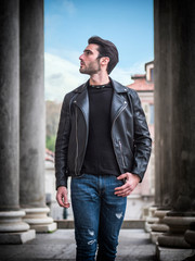Fototapeta na wymiar One handsome young man in urban setting in modern city, standing, wearing black leather jacket and jeans, looking away