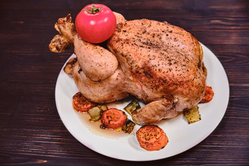 Whole grilled chicken with golden crust on a plate. Fried with tomato chicken on redwood background, mahogany. Festive dinner with baked bird.