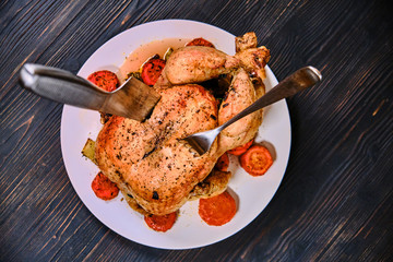 Whole grilled chicken with cutlery on a plate. Fried with carrot chicken on black wood background. Festive dinner with baked bird.