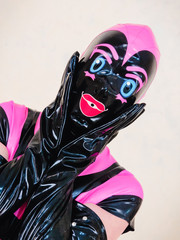 Hooded woman holding gloved hands to face in expression of shock or surprise. Pink and black rubber latex outfit with blue eyes and big red lips. Bdsm or freaky Halloween costume.