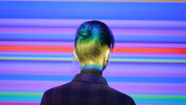 Back view of young woman looking around at modern immersive exhibition or club event with changing multi color projector light illumination. Digital art, technology and entertainment concept