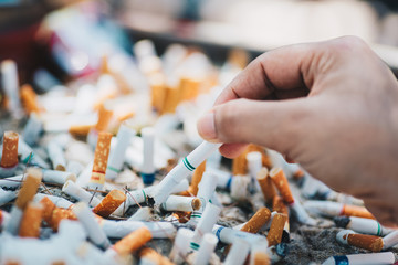 Hand putting the cigarette on an ashtray with cigarette butts stuck in ash, close up selective focus.