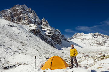 Tourist man stands near a yellow tent in the mountains in winter
