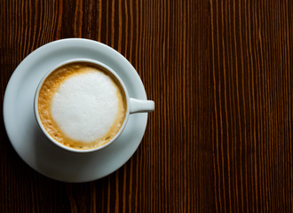 Cup of coffee with milk crema (view from above). White cup of cappuccino stands on background of brown wooden table.