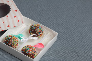 Cake Pops in chocolate with colored sprinkles. Decorated with a ribbon bow. They lie in a gift box, in the lid of which there is a transparent window.  On a gray background.