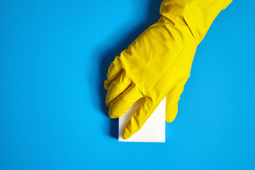 Hand in yellow protective glove with white melamine household sponge on blue background. Means for cleaning surfaces from dirt and stains