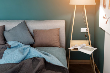 Close up of a bed with colored pillows, bedspread and a cozy lamp. Magazine on the table, evening reading. Loft interior, minimalist scandinavian style.