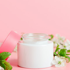 Face cream in white jar and pink cover on a pink background with white flowers. Concept natural cosmetics, organic beauty.