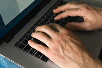 a man in jeans and a t-shirt sits on the couch and prints on a laptop. men's hands, print on the computer, close-up