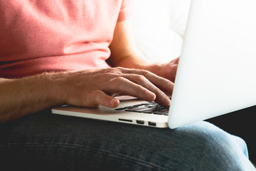 a man in jeans and a t-shirt sits on the couch and prints on a laptop. men's hands, print on the computer, close-up