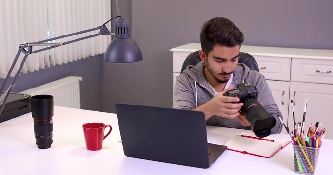 Concentrated young male photographer with camera and laptop computer sitting on table at studio and sharing his photos to social media