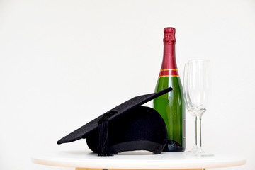 Graduation cap, champagne bottle and glasses on table with a white wall in the background