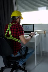 Construction Worker is sitting at his desk at the computer, looking at building plans, counting and...