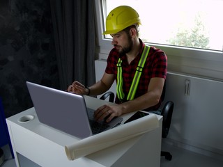 Construction Worker is sitting at his desk at the computer, looking at building plans, counting and...