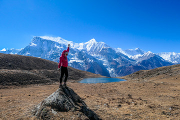 A girl wearing pink jacket stands at the rock and puts her hand up in the air. She is happy and free. In the back there is an Ice Lake and Annapurna Chain. High Himalayan mountains. Barren grass. 